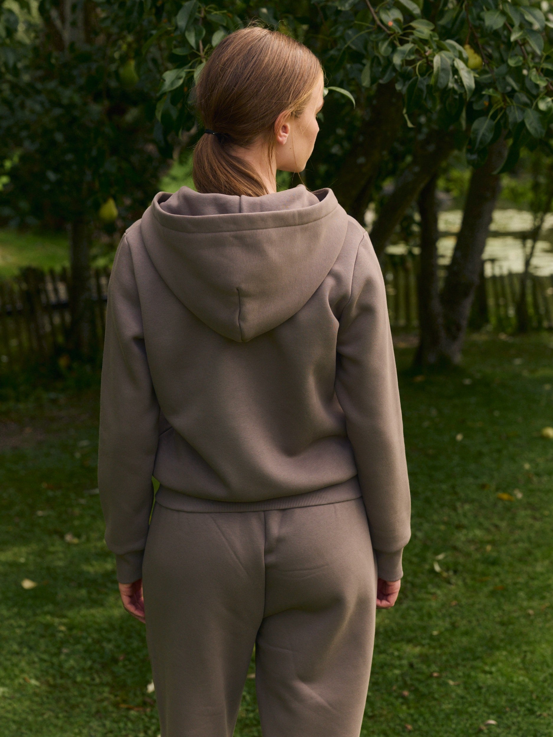 Woman seen from behind wearing a matching walnut brown hoodie and sweatpants in a garden.